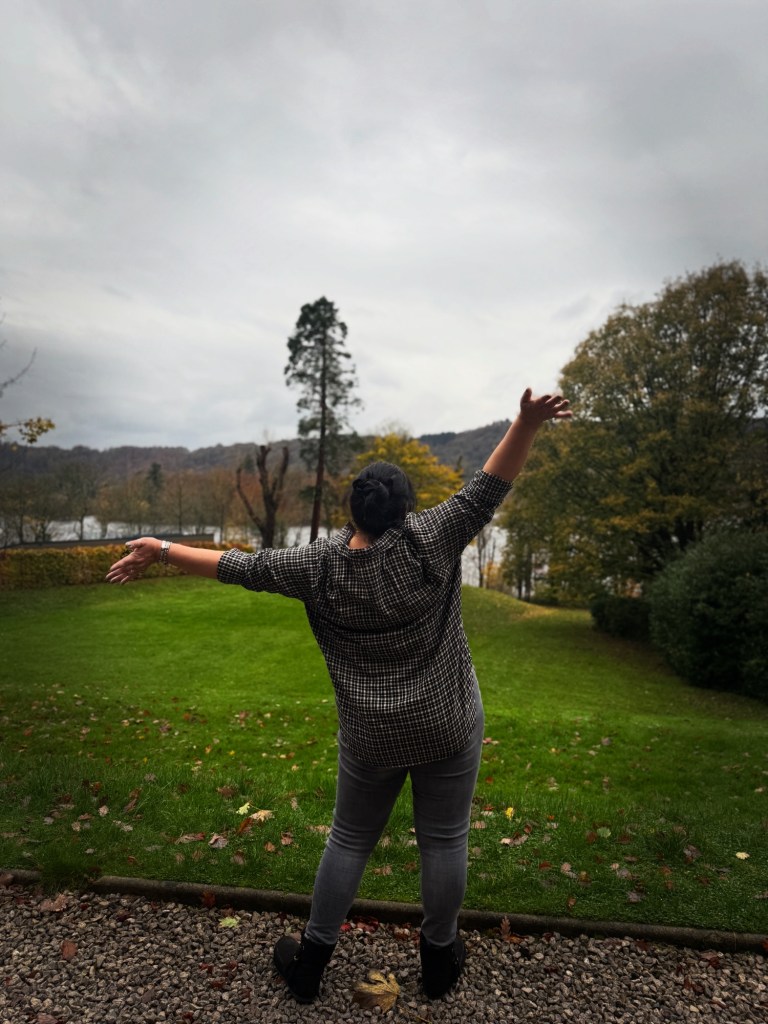 Woman standing outdoors on grass with her back to the camera, arms raised and open, overlooking trees and a lake. Dressed casually, the scene conveys ease, joy, and perspective, reflecting a grounded relationship with work, life, and alignment.