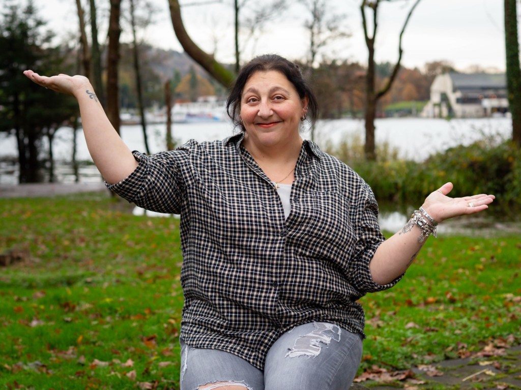Woman sitting outdoors by a lake, smiling with arms open in a confident, welcoming gesture. Dressed casually in a checked shirt and jeans, she appears grounded and approachable, reflecting calm guidance and practical support for entrepreneurs seeking direction.
