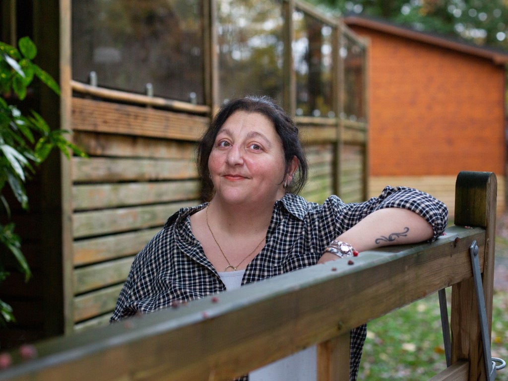 Portrait of Elena leaning on a wooden gate outdoors, looking directly at the camera with a calm, self-assured expression. Dressed casually in a checked shirt, she appears grounded and approachable, reflecting her role as a straight-talking Human Design guide and business strategist.